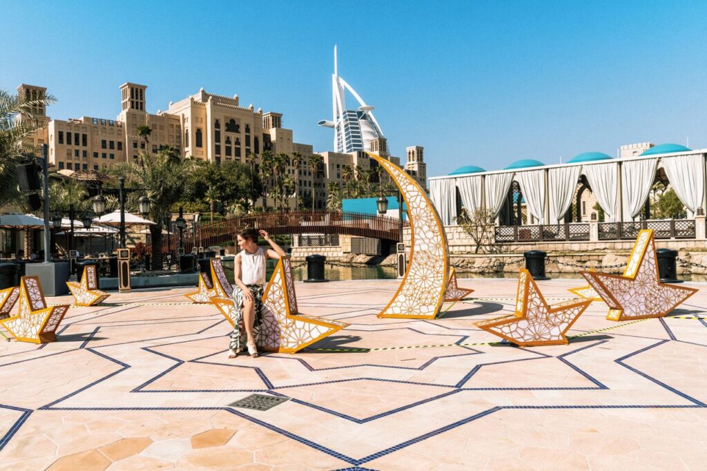 A woman relaxes by an artistic installation with the iconic Burj Al Arab in the background, set in Dubai's luxurious outdoor setting.